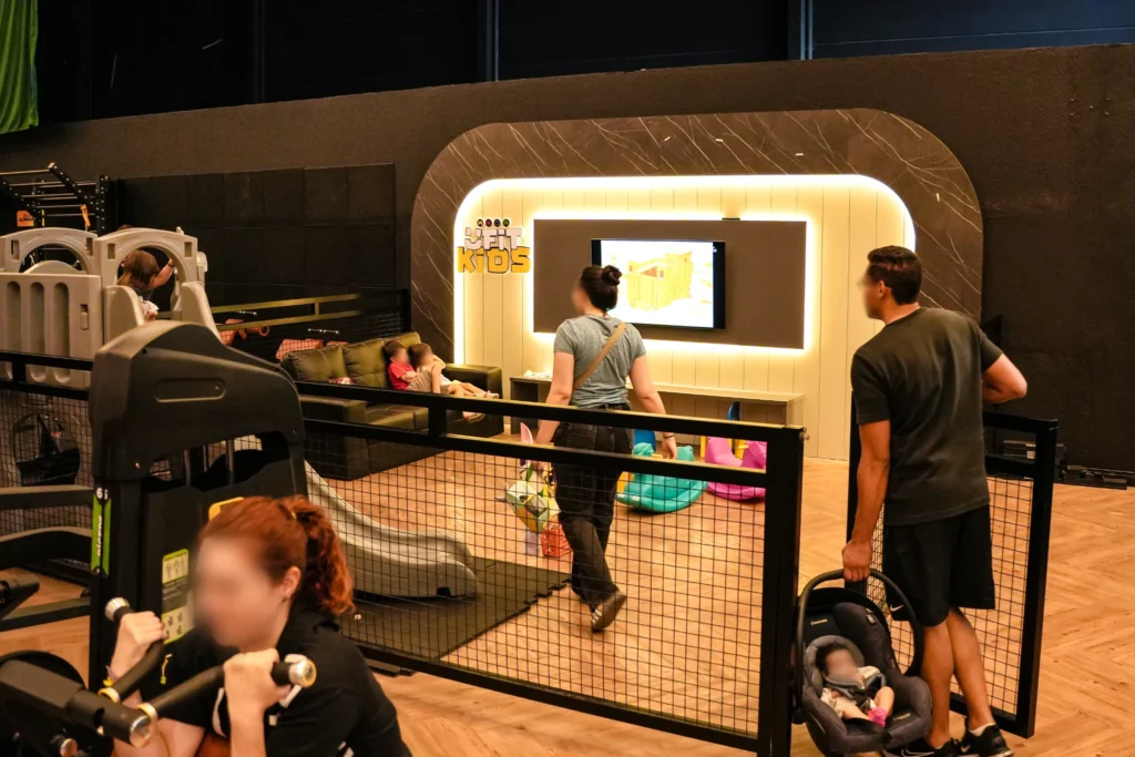 Área kids da UFit dentro do salão, com brinquedos, sofá e TV, enquanto pais treinam ao lado